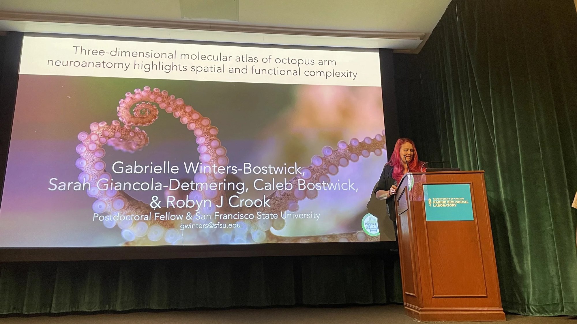 Gabrielle Winters Bostwick presenting at a lectern beside a large slide titled Three-dimensional molecular atlas of octopus arm neuroanatomy